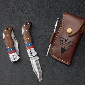 May include: Two folding knives with wooden handles and patterned blades, alongside a brown leather case. The knives have blue and red accents and the text "Name or Text". The case features a mountain graphic and the same text.