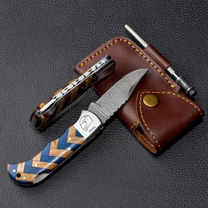 May include: A folding pocket knife with a Damascus steel blade and a handle featuring a blue and brown herringbone wood pattern. The bolster is engraved with a helmet icon and the name Justin. Includes a brown leather sheath with a metal tool.