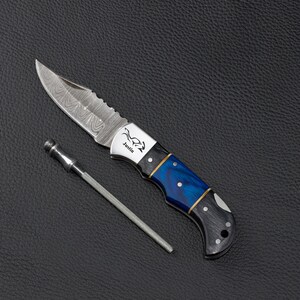 May include: A folding pocket knife with a Damascus steel blade featuring a wavy pattern, a silver bolster engraved with the name Justin, and a handle with blue and black marbled sections. A thin, metallic sharpening rod lies beside the knife.