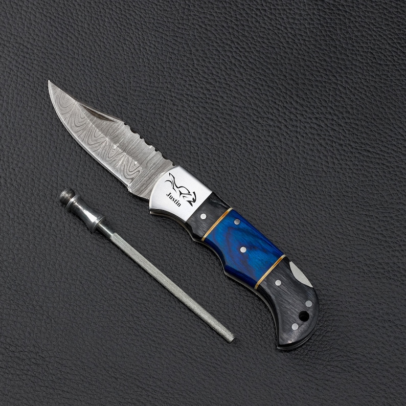 May include: A folding pocket knife with a Damascus steel blade featuring a wavy pattern, a silver bolster engraved with the name Justin, and a handle with blue and black marbled sections. A thin, metallic sharpening rod lies beside the knife.