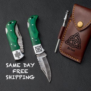 May include: Two folding knives with green handles and silver blades are displayed next to a brown leather case. The knives have decorative white accents and Celtic knot designs. The case has a Celtic knot design and the name "Brian" printed on it.