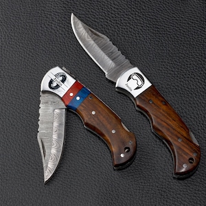 May include: Two folding pocket knives with Damascus steel blades and wooden handles. One knife is open, featuring a silver bolster with a graphic of goggles and the name Justin. The other is partially folded, with a red and blue accent and a tire graphic.