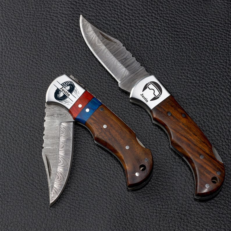 May include: Two folding pocket knives with Damascus steel blades and wooden handles. One knife is open, featuring a silver bolster with a graphic of goggles and the name Justin. The other is partially folded, with a red and blue accent and a tire graphic.