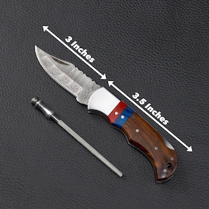 May include: A folding knife with a patterned Damascus steel blade, a silver bolster, and a handle featuring red, blue, and brown wood segments. The blade measures 3 inches and the handle measures 3.5 inches. A thin metal sharpening rod is included.