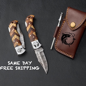 May include: Two folding pocket knives with wooden handles and a Damascus steel blade. The knives have a fish design engraved on the handle. The knives are displayed on a brown leather surface with a brown leather sheath.