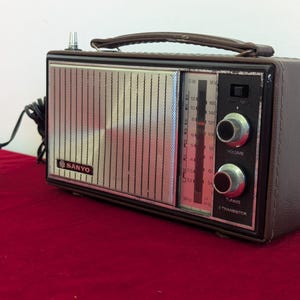 May include: A vintage Sanyo transistor radio in a brown leatherette case with a silver speaker grill. It has tuning and volume knobs, and a carrying handle. The radio is plugged into a black power adapter.