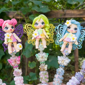May include: Three decorative pens featuring sculpted fairy figures with colorful hair, glittery wings, and floral accents. Each fairy wears a pastel dress and is mounted on a handle adorned with stacked beads, butterfly charms, and floral details.