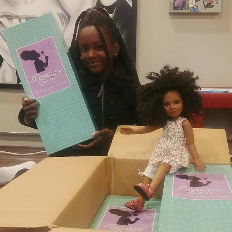 May include: A doll with dark curly hair wearing a white floral sleeveless top and white trousers, sitting in a cardboard box. The box features a light blue label with the text Herstory Doll and a silhouette graphic of a person holding a flower.