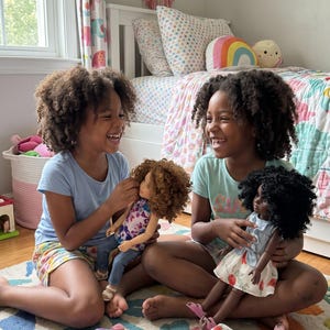 May include: Three dolls with textured hair and detailed outfits. One doll has light brown hair and wears a patterned top with blue jeans. Two dolls have black hair and wear light blue tops with floral skirts. The dolls feature articulated limbs.