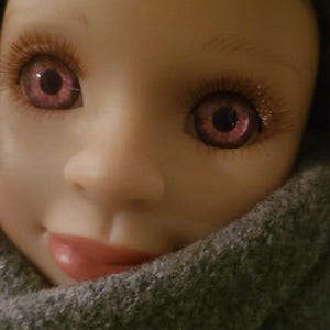 May include: A doll with a smooth, light-toned face, pinkish-purple eyes, and long brown eyelashes. The doll has brown hair and is wearing a thick, textured grey coat with a high collar that partially covers the lower portion of the face.