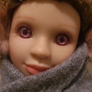 May include: A doll with curly auburn hair, light skin, and purple eyes. The doll wears a grey wool-textured coat with a single brown button and pink fuzzy earmuffs. The facial features include defined eyebrows, long eyelashes, and soft pink lips.