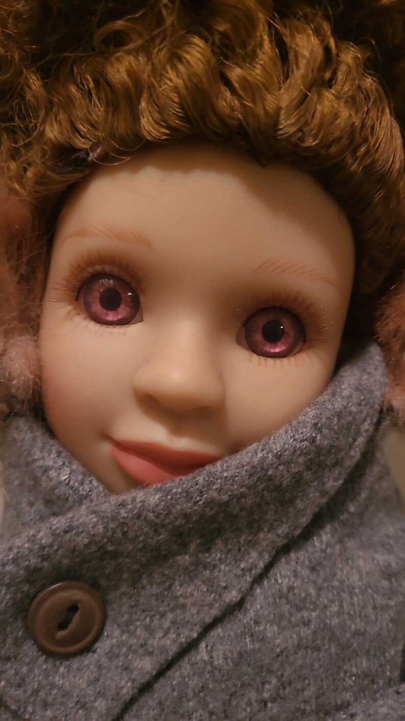 May include: A doll with curly auburn hair, light skin, and purple eyes. The doll wears a grey wool-textured coat with a single brown button and pink fuzzy earmuffs. The facial features include defined eyebrows, long eyelashes, and soft pink lips.