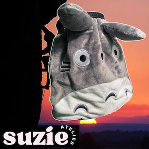 Peut inclure: Un sac en tissu gris en forme de requin de dessin animé avec de grands yeux et des oreilles. Le sac a une fermeture à cordon noire et le mot "suzie" est imprimé en bas à gauche.