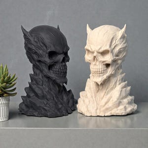 May include: Two skull sculptures, one black and one off-white, with intricate details. Each skull has a horned crown and a textured, stylized design. The sculptures are displayed on a gray surface, with a small potted plant visible on the left.