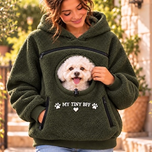 May include: An olive green sherpa fleece hoodie featuring a large front pouch with a circular opening. A small white dog is visible inside the pouch. The text "MY TINY BFF" is printed in white above two paw prints and a heart icon.