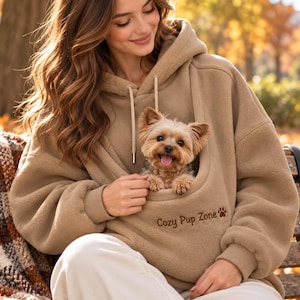 May include: A tan fleece hoodie featuring a large front pouch designed for carrying a small dog. The pouch area is embroidered with the text Cozy Pup Zone and a small paw print icon. The hoodie includes a hood with matching tan drawstrings.