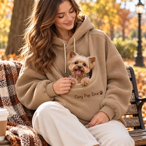 May include: A tan fleece hoodie featuring a large front pouch pocket with a small dog inside. The text "Cozy Pup Zone" and a paw print icon are embroidered in brown on the front of the pouch. The hoodie has long sleeves and a hood.