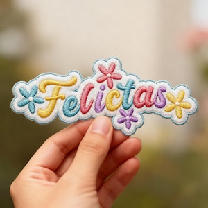 May include: A colourful embroidered patch spelling "Felicitas". The patch has a white border and is decorated with embroidered flowers in shades of pink, blue, yellow, and purple. The patch is held in a hand.