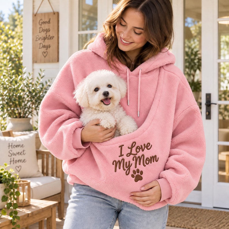May include: A light pink, long-sleeved, hooded fleece sweatshirt featuring a large front pouch containing a small white dog. The sweatshirt has the dark brown embroidered text "I Love My Mom" above a small brown paw print graphic.