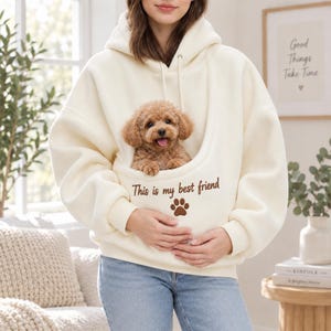 May include: An off-white hooded sweatshirt featuring a large front pouch pocket containing a small brown curly-haired puppy. The text "This is my best friend" is embroidered in brown above a brown paw print icon on the front of the pouch.