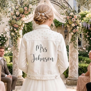 May include: A cream denim jacket adorned with pearls and the name "Mrs Johnson" in black script. The jacket is part of a bridal outfit, worn over a white tulle skirt. The background shows wedding decorations.