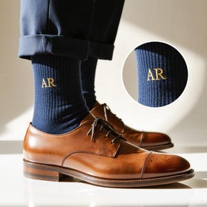 Puede incluir: Zapatos de vestir de cuero marrón con calcetines azul marino bordados con las letras doradas "AR". Los zapatos tienen un diseño clásico con puntera y cierre de cordones. Los calcetines son visibles por encima de los zapatos.