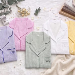 Pode incluir: Cinco camisas de pijama listradas em várias cores: lavanda, rosa, verde, branco e amarelo. Cada camisa tem um bolso no peito e iniciais bordadas. As camisas estão sobre uma superfície branca.
