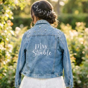 Pode incluir: Casaco de ganga azul claro com detalhes de pérolas e o texto branco "Mrs. Struble". A data "06.24.2025" também é visível. O casaco é usado sobre um vestido branco, com um acessório de cabelo floral.