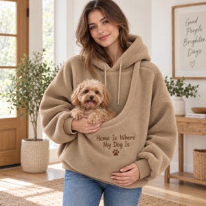 May include: A tan sherpa fleece hoodie featuring a large front pouch designed for carrying a small dog. The hoodie includes a drawstring hood, long sleeves with ribbed cuffs, and the embroidered text Home Is Where My Dog Is with a paw print.