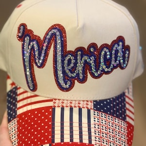 May include: A cream-colored baseball cap featuring the word 'Merica' in cursive script on the front panel. The lettering is embellished with silver rhinestones and outlined in red and blue glitter. The brim has a patchwork pattern of stars and stripes.