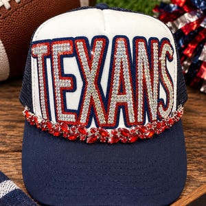 May include: A navy blue and white trucker hat featuring the word TEXANS in large, iridescent rhinestone-encrusted letters with a red border. A decorative band of marquise-cut red crystals sits horizontally across the base of the front panel.