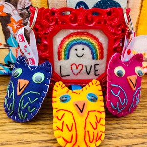 May include: Three felt owl keychains with embroidered details. One owl is blue with green accents, one is yellow with red accents, and one is pink with purple accents. The owls are hanging from a red picture frame with a rainbow and the word "LOVE" embroidered on it.