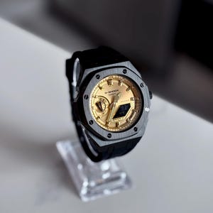 Könnte beinhalten: Eine schwarze und goldene G-Shock Casio-Uhr mit einem schwarzen Kautschukarmband. Die Uhr hat ein goldenes Zifferblatt mit schwarzen Akzenten und wird auf einem transparenten Ständer präsentiert. Das Gehäuse ist quadratisch und hat eine Digitalanzeige.