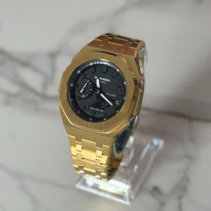 Könnte beinhalten: Eine goldfarbene Armbanduhr mit schwarzem Zifferblatt und passendem goldenem Armband. Die Uhr wird auf einem transparenten Ständer präsentiert, der ihr aufwendiges Design und ihre metallische Oberfläche hervorhebt. Klassische analoge Anzeige.