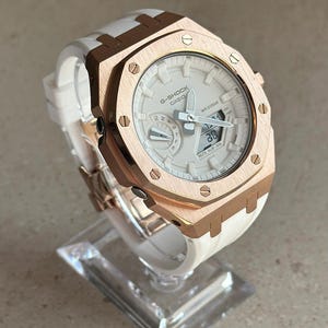 May include: A rose gold G-Shock watch with a white face and white silicone band. The watch has a digital display and a date window. The watch is water resistant to 20 bar.