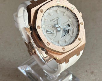 Peut inclure: Une montre G-Shock en or rose avec un cadran blanc et un bracelet en silicone blanc. La montre possède un affichage numérique et une fenêtre de date. La montre est étanche à 20 bars.