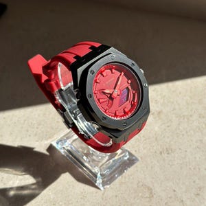 Op de afbeelding: Een rode G-Shock horloge met een zwarte bezel en een rode band. Het horloge wordt getoond op een heldere, schuine standaard. De wijzerplaat is rood met zwarte accenten.