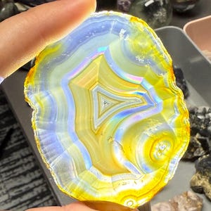 May include: A translucent, polished agate slice with concentric bands of yellow, blue, and white. The center features a geometric pattern. The stone is held up to the light, revealing its internal structure and colors. Other gemstones are visible in the background.