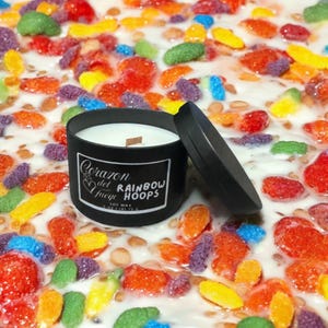 May include: A black candle jar with a wooden wick. The label reads "Corazon del Fuego" and "Rainbow Hoops" with a heart and a soy wax logo. The candle is sitting on a white surface with colorful cereal pieces.