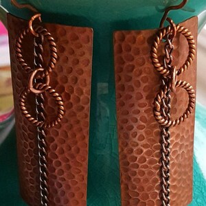 May include: A pair of copper earrings with a hammered texture. Each earring features a rectangular copper piece with a chain and three copper rings hanging from it.