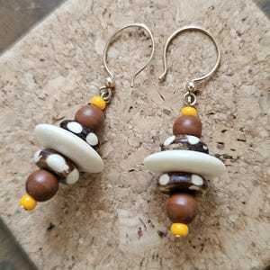 May include: A pair of dangle earrings with brown, white, and yellow beads. The beads are stacked on top of each other and are connected by a silver wire.