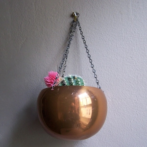May include: A polished copper-colored spherical hanging planter suspended by a dark metal chain. The bowl-shaped container holds a small green cactus with white spines and a bright pink flower bloom, reflecting light on its smooth surface.