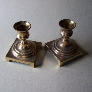 May include: Two brass candle holders. Each has a square base with a rounded top and a cup-shaped holder. The candle holders are made of a shiny, golden-colored metal and are placed on a light gray surface.