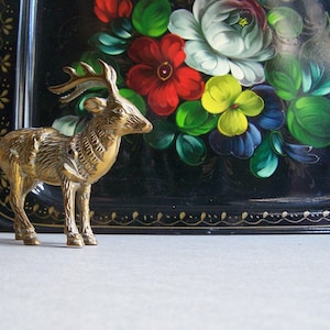 May include: A gold-colored metal deer figurine with a detailed design. The figurine is standing on a black tray with a painted floral design featuring red, yellow, and blue flowers.