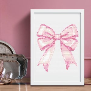 Könnte beinhalten: Ein gerahmtes Kreuzstich-Kunstwerk mit einer großen rosa Schleife. Die Schleife ist mit verschiedenen Rosatönen detailliert, was ihr ein strukturiertes Aussehen verleiht. Das Kunstwerk befindet sich vor einem weißen Hintergrund in einem weißen Rahmen, geeignet für die Inneneinrichtung.