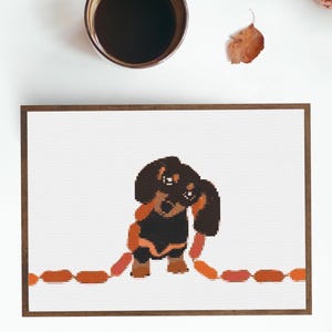 Op de afbeelding: Een ingelijst kruissteekontwerp met een teckel met een ketting van worstjes. De hond is zwart en bruin, met oranje worstjes. Een kopje koffie en een blad staan op de achtergrond.