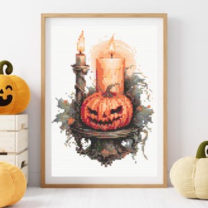 May include: A framed cross-stitch artwork with a Halloween theme. The image shows a carved pumpkin with a candle, a tall candle, and foliage. The artwork is in a wooden frame and is surrounded by decorative pumpkins.
