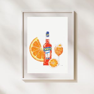 May include: Cross-stitch art print featuring a bottle of Aperol, a glass of Aperol Spritz, and orange slices. The bottle is red with a blue and yellow label. The drink and orange slices are orange. The artwork is framed in a white frame.