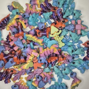 May include: A pile of small, multi-coloured, jointed animal figures in shades of blue, purple, orange, yellow, and green. The figurines look 3D-printed and are in a white bowl. The toys have a fantasy theme.