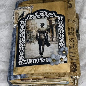 Op de afbeelding: Handgemaakt dagboek in vintage stijl. De omslag toont een zwart-wit afbeelding van een man met hoge hoed, omlijst in zwart. Versierd met papier, stof en kleine, lichtblauwe bloemen. Een uniek, handgemaakt item.